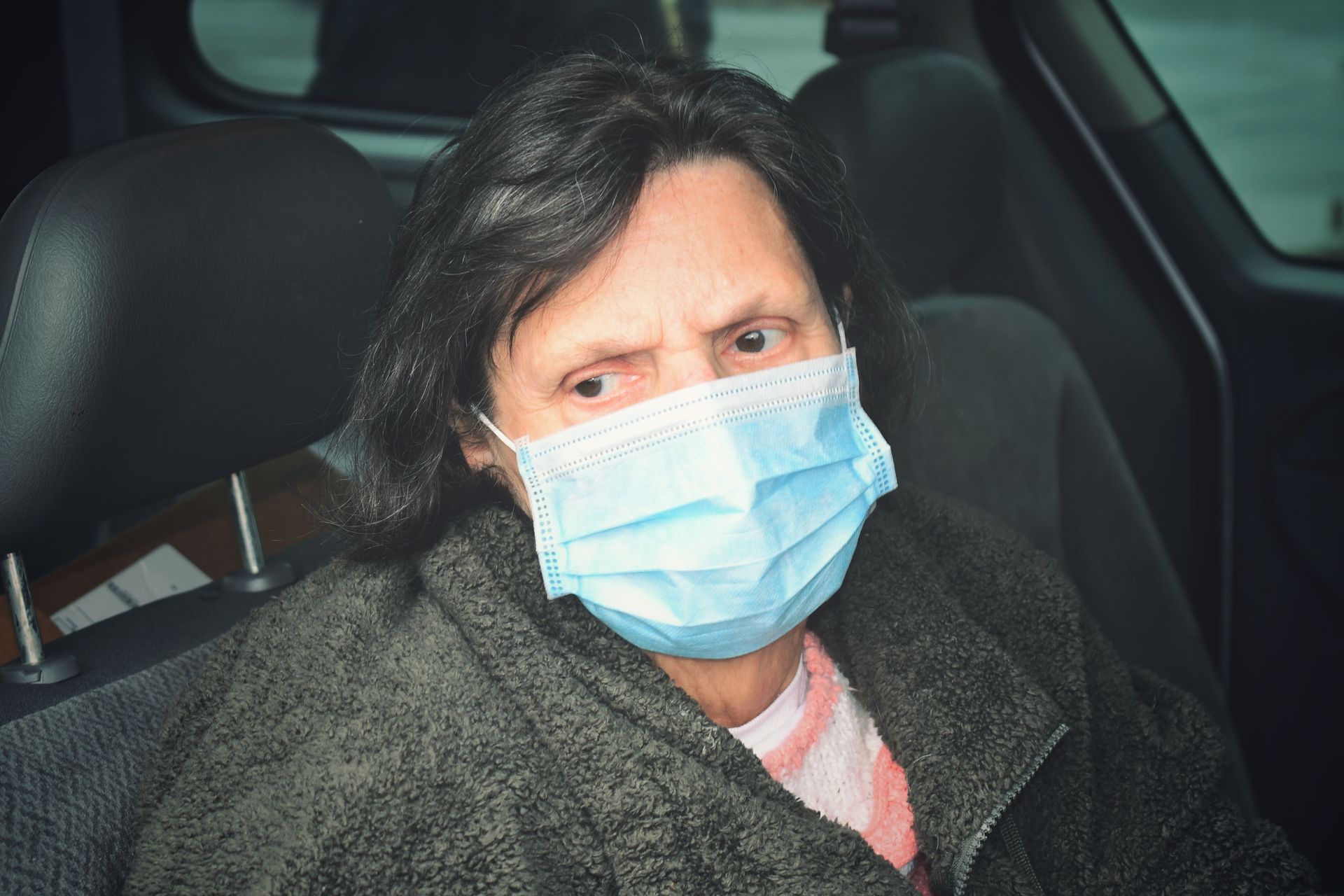 Senior woman wearing a face mask sitting inside a car, looking outside with concern.